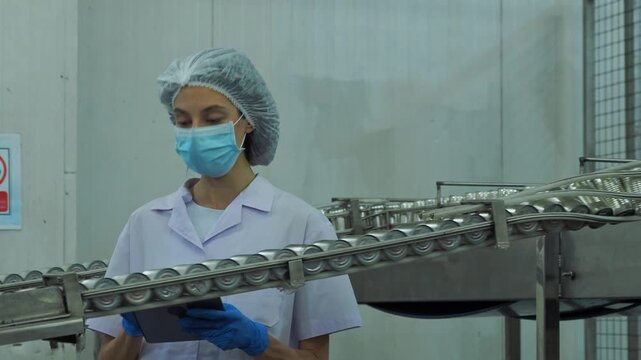 focused female quality specialist use digital tablet while inspect row metal can moving along automated conveyor track conducting compliance audits production monitor modern food packaging facility