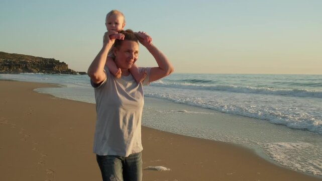 Happy mother with baby on neck walking on the sandy beach on summer vacation. Caring mother and her toddler child in nature in summer by sea while sunset.  Happy motherhood concept. Active leisure