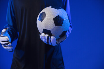 Fototapeta premium African American man holding black-white soccer ball at chest in blue studio, dark jersey, gloves
