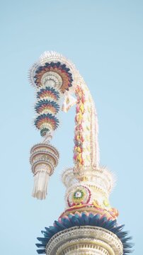Bali Hindu Festival Bamboo Pole with Cascading Multicolor Disc Ornaments and Deep Teal Feather Base Against Pale Blue Sky