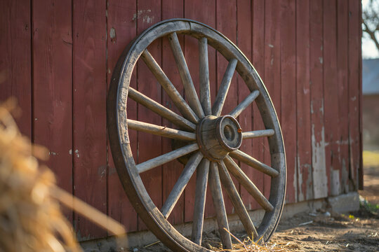A weathered wooden wagon wheel leaning against a rustic red barn wall on a farm with a blurred hay bale in the foreground during the day