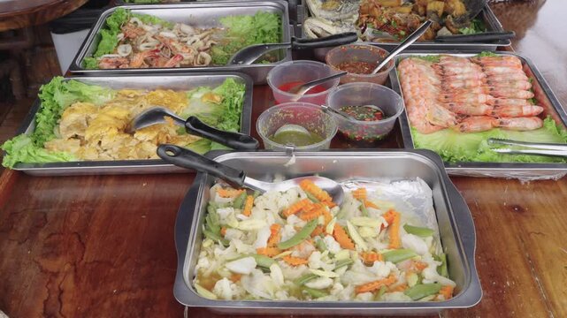 Assorted Thai seafood buffet with grilled shrimp, stir-fried vegetables, squid salad, and spicy dipping sauces served in metal trays on a wooden table. Professional catering food display.