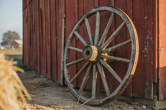 An old wooden wagon wheel with iron rim leaning against the side of a weathered red wooden barn on a farm during a bright sunny afternoon