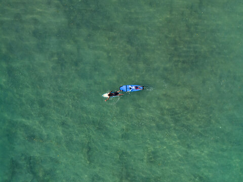 Aerial view of surfing training at Nusa Dua Beach with two surfers on blue surfboards in clear turquoise water in South Kuta, Bali, Indonesia.