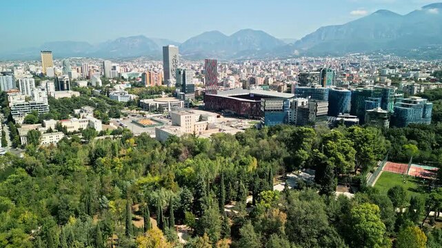 Albania, Tirana 06 09 2025: Forward dolly drone video over Grand Park trees toward Arena Kombetare, Marriott, Mother Teresa Square and city center skyline backed by mountains.