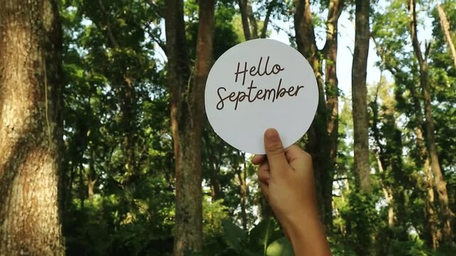 A person holding a circular sign that says Hello September in a forest.