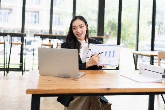 A smiling Asian businesswoman holding a financial chart and pointing at it while having a video conference call on her laptop in a modern office.