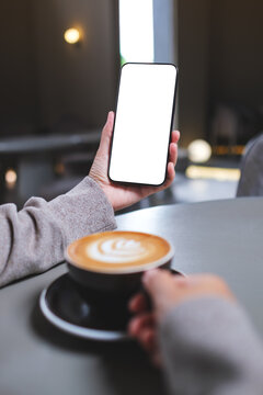 Vertical mockup image of a woman holding mobile phone with blank desktop screen while drinking coffee in cafe