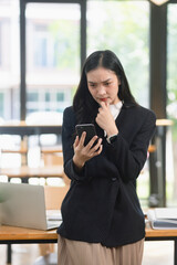 Fototapeta premium A serious Asian businesswoman looking confused and thinking while holding a smartphone in the office.