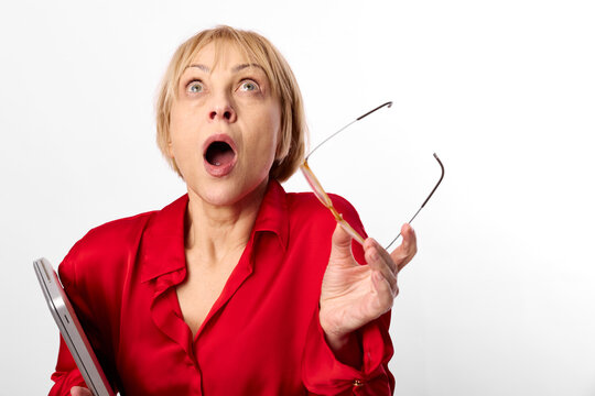 Surprised shocked woman in red blouse holding eyeglasses, portrait with wide open mouth, looking upward, studio white background, attentive startled expression closeup