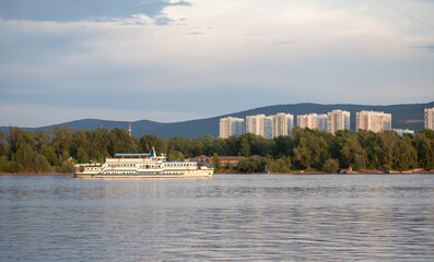 Naklejka premium Riverboat navigates calm waters with a city skyline and mountains visible in the background, creating a tranquil landscape at sunset