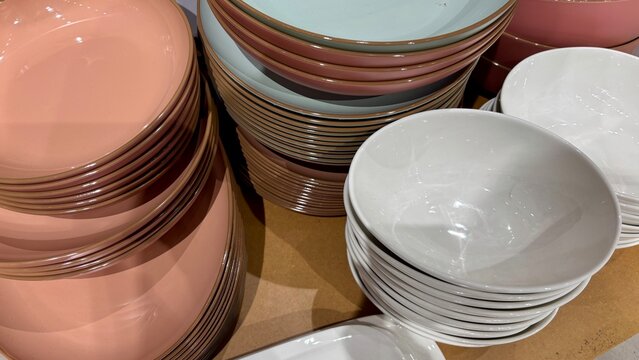 Closeup of stacked ceramic plates and bowls in pink, white and neutral tones displayed in a homeware store, showing varied shapes, sizes and glossy finishes