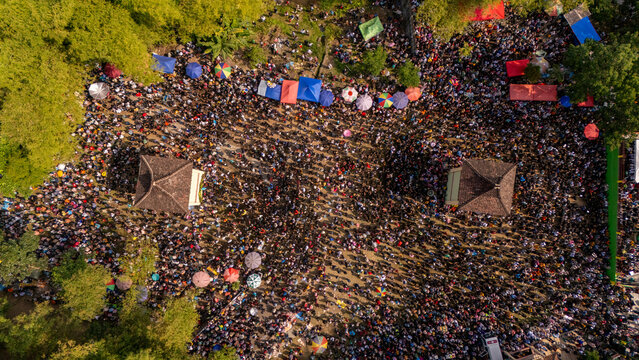 Aerial view of a massive crowd gathered in an open field with colorful umbrellas and two traditional structures in Klaten, Central Java, Indonesia.
