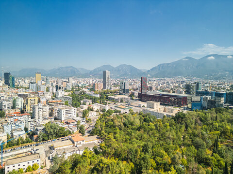 Albania, Tirana 06 09 2025: Wide angle aerial drone photo of Mother Teresa Square, Arena stadium, Marriott and university past park trees with city center skyline and cloudy mountains.