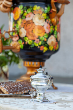 Traditional Russian samovar and a silver teapot placed on a decorative table with floral patterns, accompanied by baked goods in the background