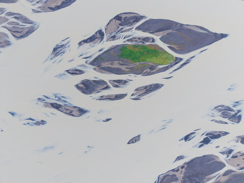 Aerial view of a braided river system with milky glacial water flowing around dark sediment islands and a patch of green vegetation in Iceland.