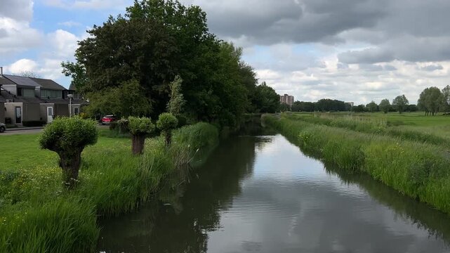 Amsterdan Forest, Suburban canal panorama with reflective water, gentle ripples, mown lawn, reed grasses, modern townhouses, parked cars, pollard trees, and overcast sky above quiet residential