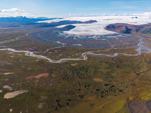 Aerial view of a vast glacier tongue flowing into a braided river system across a mossy volcanic landscape under a bright sky in Iceland.