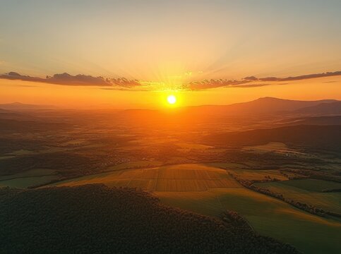 radiant golden sunset over patchwork fields and forested hills with sun rays through thin clouds and distant mountains creating a calm, peaceful atmosphere