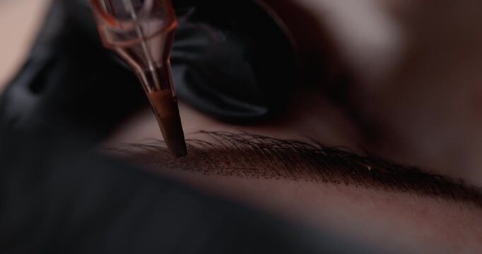 Handheld tracking shot of microblading artist shading eyebrow hairs on client's face in beauty studio close up macro.