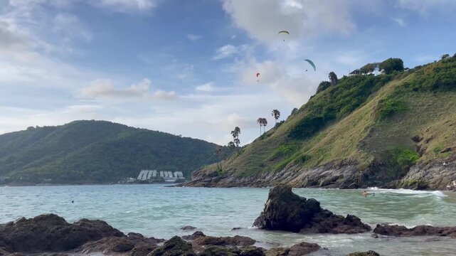Paragliders fly above a scenic tropical bay in Phuket, Thailand, with turquoise water, rocky coastline and lush green hills under a partly cloudy blue sky