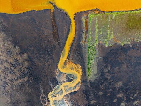 Aerial view of a vibrant yellow river delta winding through dark volcanic terrain and patches of green moss in Iceland.