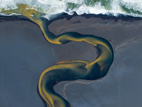 Aerial view of a winding river with golden sediment flowing across a dark black sand beach into the churning white waves of the ocean in Iceland.