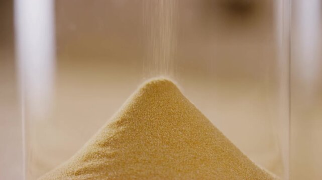 Slow motion static close up of sand grains pouring and accumulating into pile inside hourglass on neutral background studio table