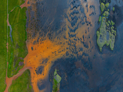 Aerial view of a river delta showcasing vibrant orange sediment patterns contrasting with dark water and lush green grassy banks in Iceland.