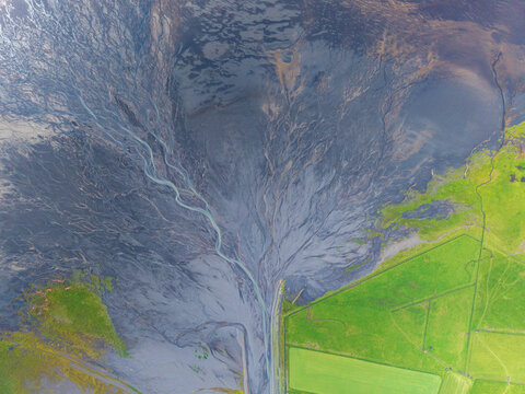 Aerial view of a braided river system with dark volcanic sediment flowing through vibrant green fields in Iceland.