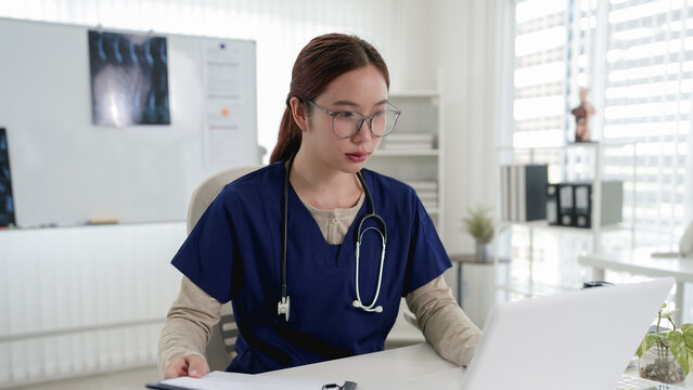 Young Asian female doctor wearing glasses and stethoscope working on laptop in modern bright medical office
