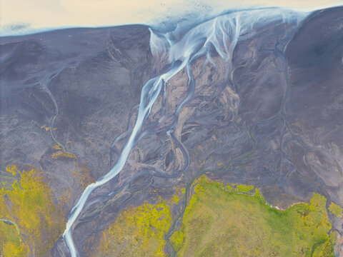 Aerial view of a glacial river delta with intricate braided channels, dark volcanic sand, and vibrant green mossy vegetation in Iceland.