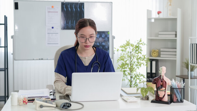 Young Asian female doctor typing on laptop during telehealth consultation in bright modern clinic office with medical equipment
