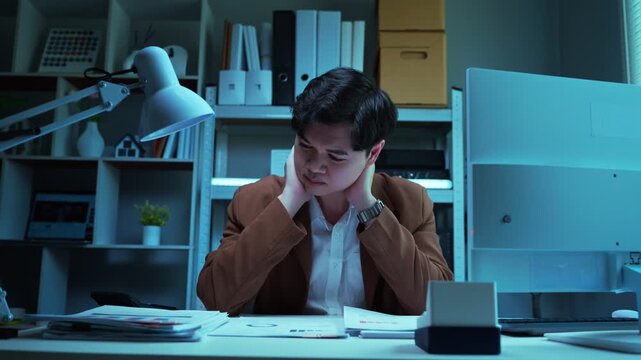 Stressed and exhausted Asian businessman holding head working late overtime at dark office