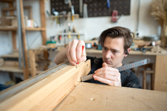 Woodshop employee working with old window frame parts at worktable in restoration workshop, preparing patch and marking drawing on board with pencil, master checking fitting for future replacement.