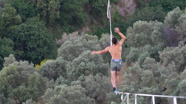 A man balances on a tightrope in the mountains. An athletic, shirtless man carefully balances and walks along a tightrope on a mountainside among green hills. Stilo, Italy. August 15, 2026.