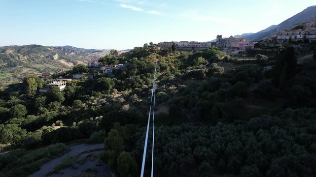 Drone footage shows a highline sling strung over a mountain valley. The highline sling and rigging are visible strung over the valley against the backdrop of a picturesque Italian hillside town.