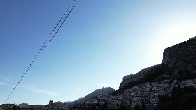 Drone footage shows a highline sling strung over a mountain valley. The highline sling and rigging are visible strung over the valley against the backdrop of a picturesque Italian hillside town.