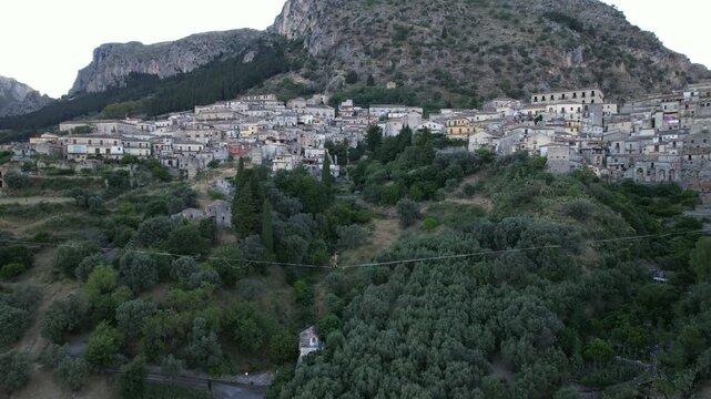 An aerial view of a tightrope walker walking above a deep valley. The tightrope walker carefully balances on the rope above a lush green canyon and olive trees. Italy. Stilo, August 15, 2026.