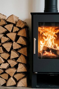 Black modern wood burning stove with bright orange flames inside. Stack of cut firewood logs rests next to heater. Cozy warm home interior detail.