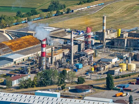 Aerial view of an industrial facility with smoking chimneys, factory buildings, and storage tanks, situated near railway tracks and agricultural fields in Hombourg, Grand Est, France.