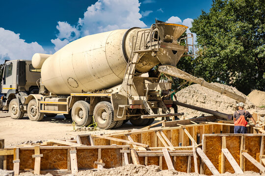 Construction workers are operating a concrete mixer at a building site. They pour cement into wooden forms. Workers are busy with their tasks in the afternoon