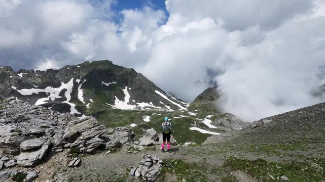 Drone tilt up shot of a lone hiker with backpack standing on a mountain pass, revealing epic peaks and massive clouds