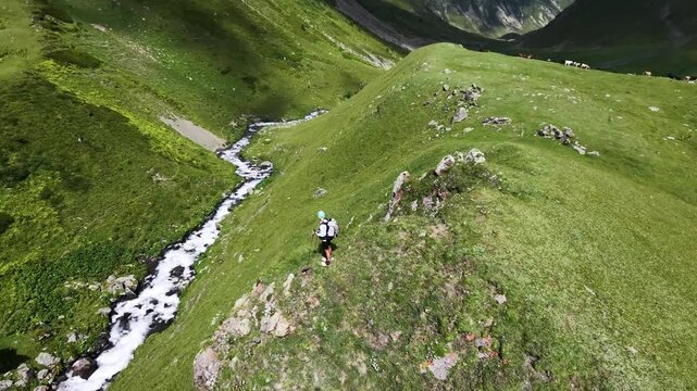 Drone shot of a tourist trekking on a green alpine slope, standing on a mountain summit in summer