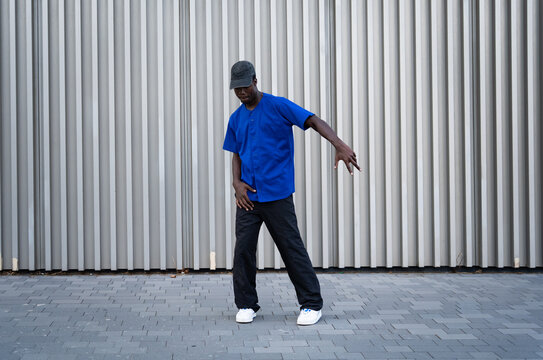 Young man wearing a blue shirt and baseball cap, performing street dance moves on a city sidewalk