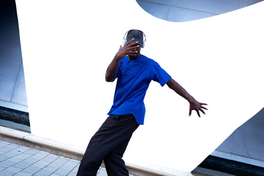 Black man wearing headphones and a blue shirt, performing a dance move in a vibrant outdoor city location