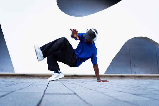 Young black man performing a breakdance move on a paved urban floor, listening to music with headphones
