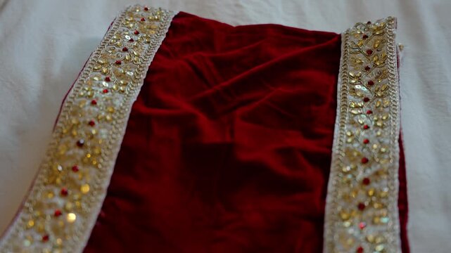 Folded red velvet groom garment with intricate gold and silver beaded embroidered borders laid on white bed sheet overhead angle