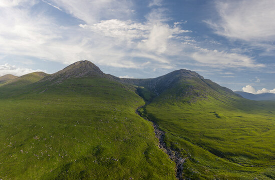 Aerial view of Glen Etive, featuring lush green mountains, a rocky stream flowing through the valley, and a bright blue sky with wispy clouds in Ballachulish, Scotland, United Kingdom.