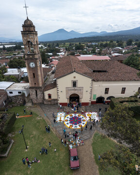 Aerial view of the intricate Patamban floral rug being arranged by people in the church courtyard, with historic architecture and mountains in Patamban, Michoacan, Mexico.